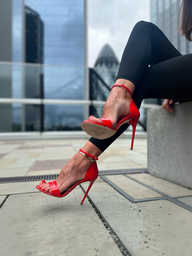 Red patent leather stiletto sandals worn by a woman sitting outdoors against a city skyline.