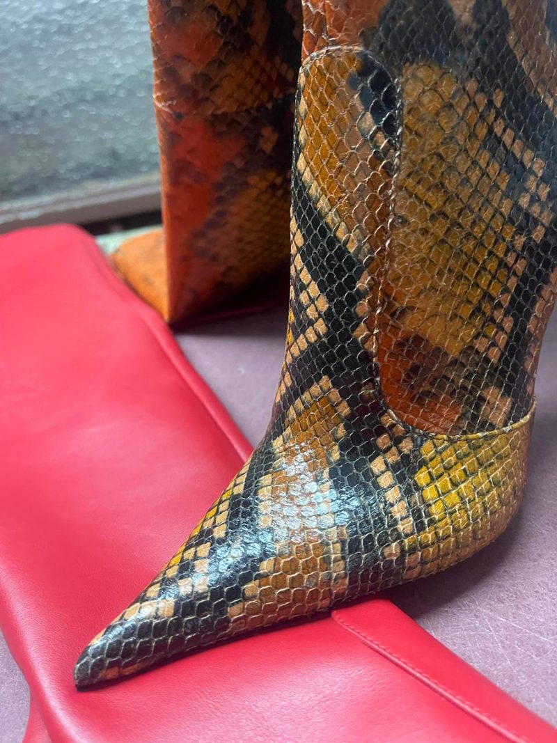 Close-up of the extreme pointy toe of a python leather boot resting on a vibrant red leather surface.