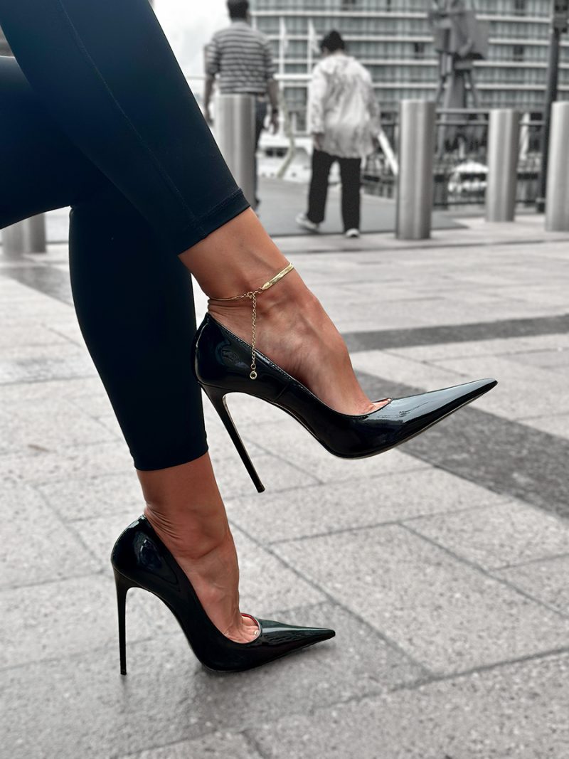 Model with a delicate gold anklet wearing 1969 LX E27 black patent leather stiletto pumps on a city pavement.
