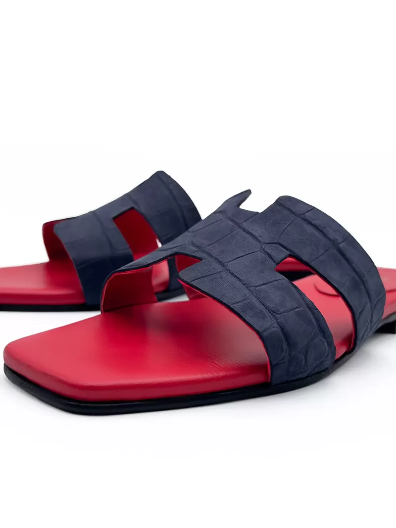 Staggered view of dark blue croc-embossed suede flat slide sandals with contrasting red footbeds.