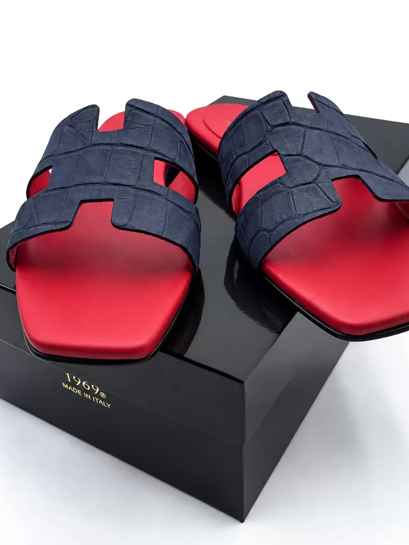 Dark blue croc-embossed suede flat slide sandals resting on a black box with gold logo.