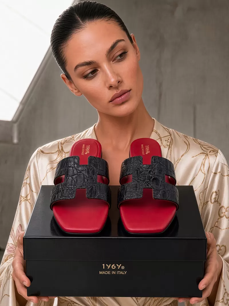 Model holding Oran black croc-embossed leather flat sandals with red footbeds on a 1969 Made in Italy shoe box.
