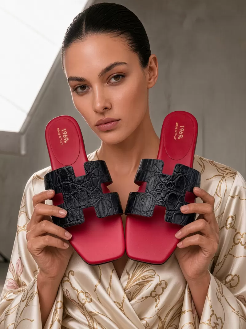 Model holding Oran black croc-embossed leather flat sandals with H-cut straps, square toes and red leather footbeds.