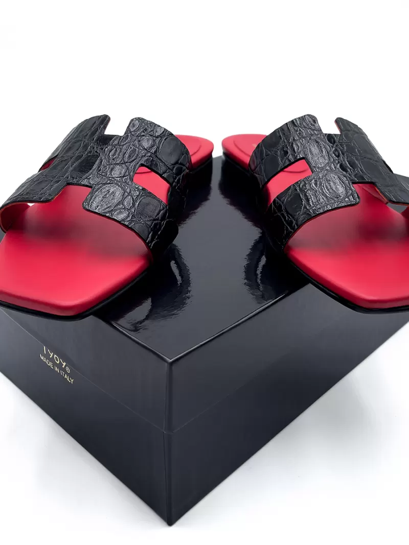 Front view of 1969 Oran black croc leather flat sandals displayed on top of a luxury black shoe box with gold branding.