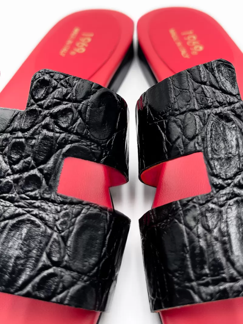Close-up of 1969 black croc-embossed leather flat sandals showing H-strap texture, red lining and gold logo.