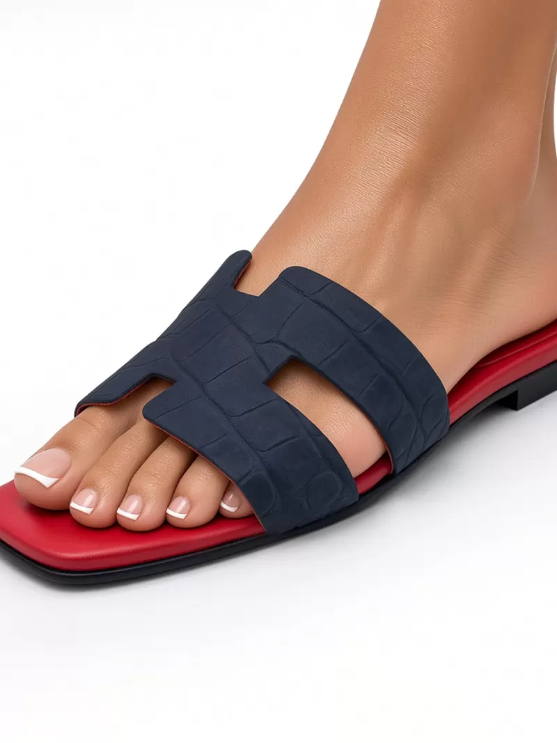 Side view of a model wearing an Oran blue croc-embossed suede leather flat sandal with H-cut strap, square toe and red footbed.