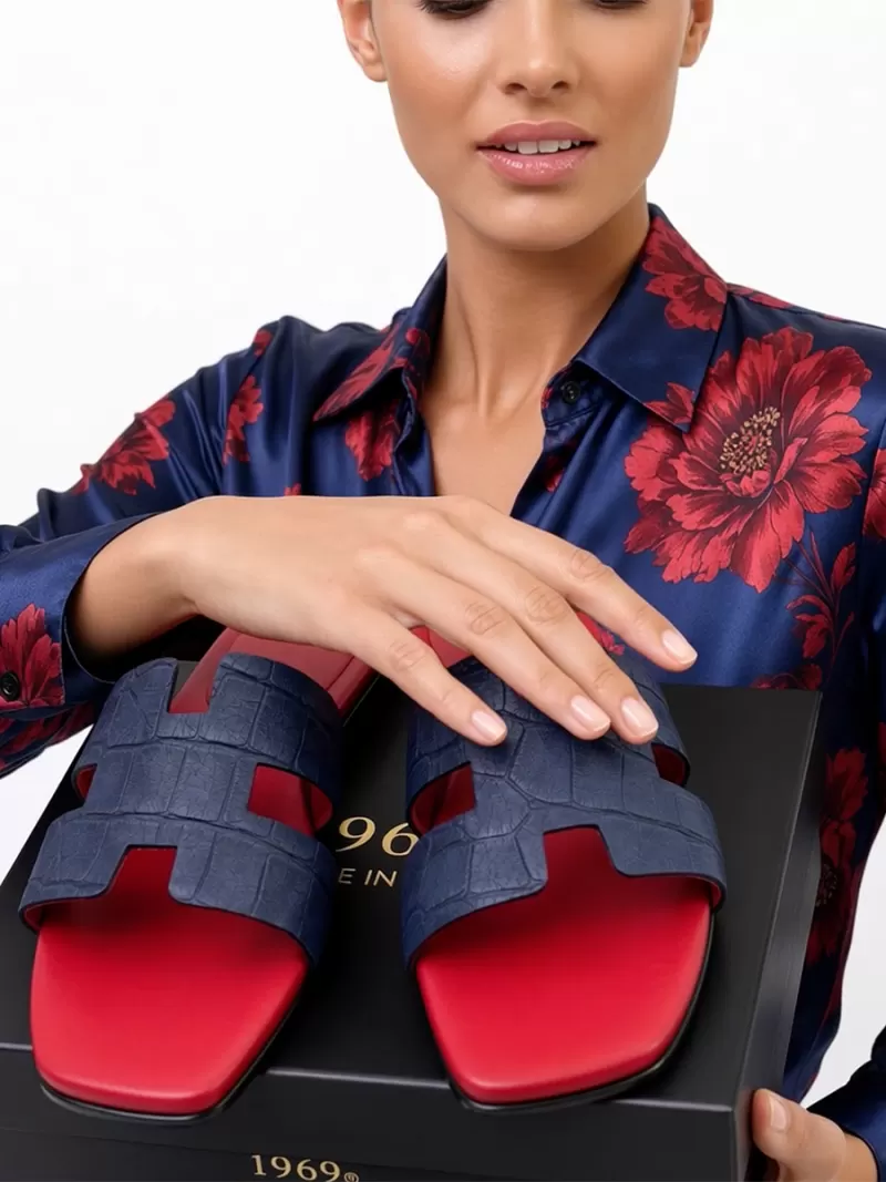 Close-up of Oran blue croc-embossed suede leather flat sandals with H-cut uppers and red footbeds on a shoe box held by a model.