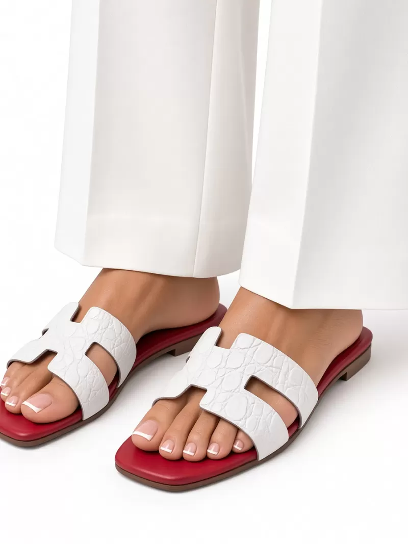 Close-up of Oran white croc-embossed leather flat sandals worn with white trousers, showing H-cut uppers and red footbeds.