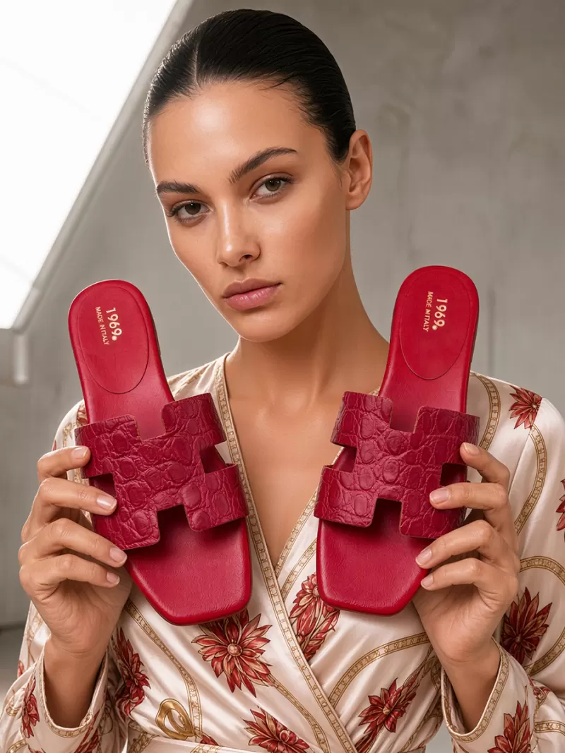 Model holding a pair of 1969 red croc-embossed leather flat slide sandals with H-cut straps and red footbeds.