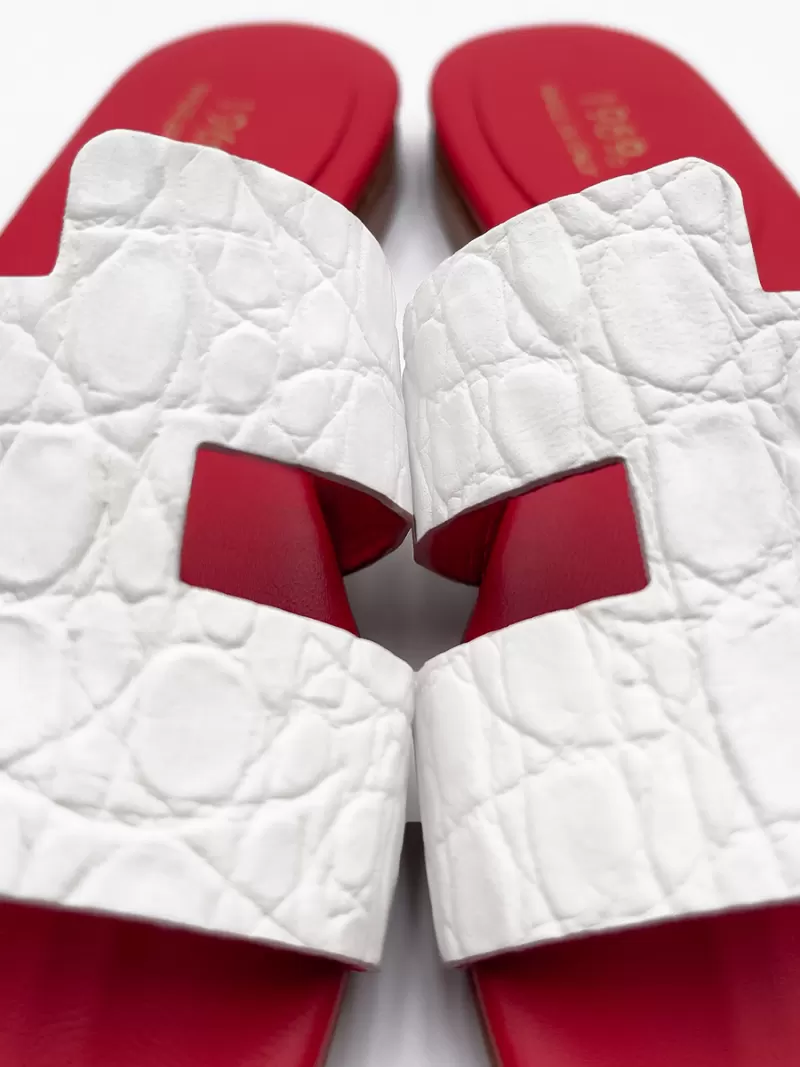 Macro shot of white croc-embossed leather H-strap flat sandals with signature red footbed.